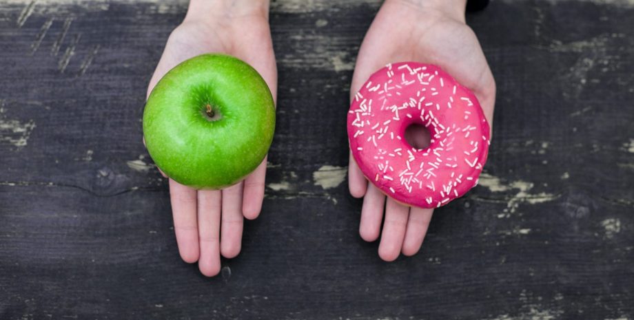 A hand holding a green apple and a hand holding a pink sprinkle doughnut